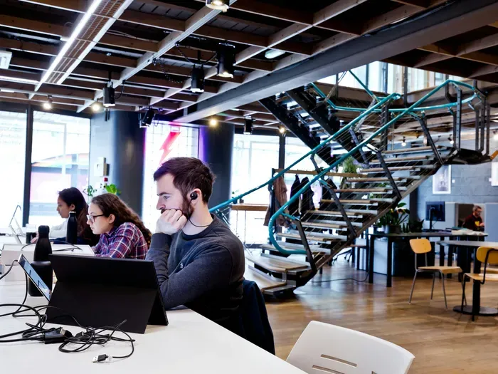 People working on laptops at shared desks in a bright coworking office with open layout and spiral staircase
